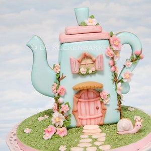 Puede incluir: Una tetera azul y rosa con forma de casa con una puerta rosa, una ventana y un techo rosa. La tetera está decorada con flores rosas y enredaderas. Un caracol está sentado en la hierba frente a la tetera.