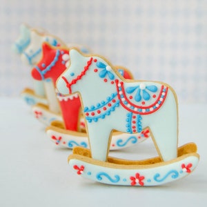 May include: Rocking horse cookies in a row, decorated with light blue, red, and white icing. The cookies have intricate details, including floral and dot patterns. The cookies are on a white surface.