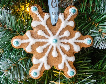 Könnte beinhalten: Ein Lebkuchen-Schneeflocken-Keks-Ornament mit weißem Zuckerguss und blauen Akzenten, das an einem hellblauen Band hängt. Das Ornament ist mit weißen Zuckerkristallen und blauen Perlen verziert. Auf einem Weihnachtsbaum.