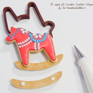 May include: A decorated red rocking horse cookie with blue and white details, next to a brown cookie cutter and a pastry bag. The cookie is on a light brown rocker base. The image includes the text "&copy; 2017 3D Cookie Cutter Shop by De Koekenbakkers."