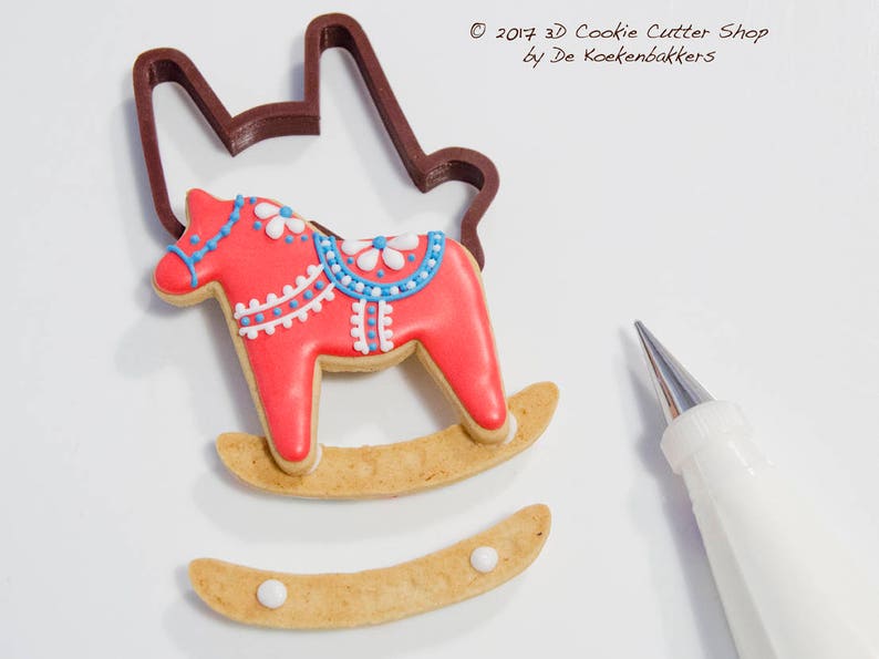 May include: A decorated red rocking horse cookie with blue and white details, next to a brown cookie cutter and a pastry bag. The cookie is on a light brown rocker base. The image includes the text "&copy; 2017 3D Cookie Cutter Shop by De Koekenbakkers."