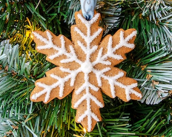 Könnte beinhalten: Ein schneeförmiger Keks-Ornament mit weißen Zuckergussdetails und einem hellblauen Band zum Aufhängen. Der Keks hat eine hellbraune Farbe und ist mit weißem Zuckerguss in einem Schneeflockenmuster verziert. Das Ornament wird auf einem Weihnachtsbaum präsentiert.