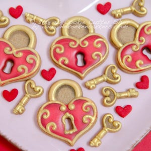 Op de afbeelding: Een set hartvormige slot- en sleutelkoekjes versierd met rode en gouden glazuur. De koekjes zijn gerangschikt op een roze bord met rode hartjes.