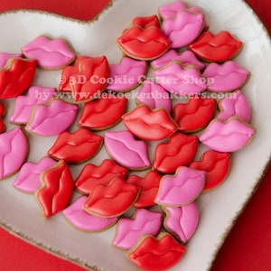 Op de afbeelding: Een hartvormige schaal gevuld met rode en roze lipvormige koekjes. De koekjes zijn versierd met glad glazuur en liggen op een witte schaal met een getextureerde rand. De achtergrond is effen rood. De tekst "© 3D Cookie Cutter Shop" is zichtbaar.
