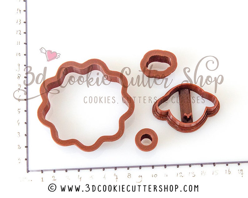 May include: A set of four brown plastic cookie cutters. The cutters are shaped like a flower, a sheep, and two circles. The cutters are on a white surface with a ruler in the background.