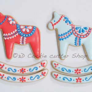 May include: Two Dala horse cookies, one red and one light blue, with intricate white and blue icing details. The cookies are decorated with floral and dot patterns. Two matching rocker base cookies are also present, featuring similar designs. The text "3D Cookie cutter Shop" is visible.