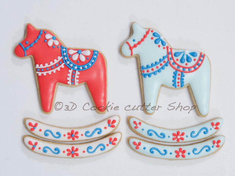 May include: Two Dala horse cookies, one red and one light blue, with intricate white and blue icing details. The cookies are decorated with floral and dot patterns. Two matching rocker base cookies are also present, featuring similar designs. The text "3D Cookie cutter Shop" is visible.