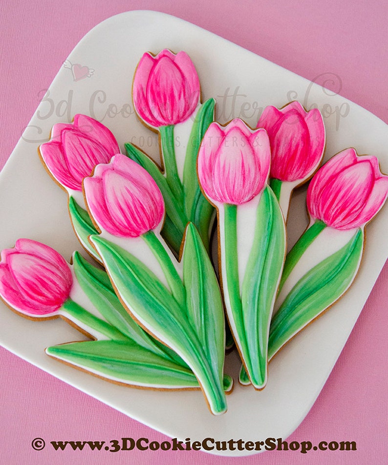 May include: A bouquet of pink and green sugar cookies decorated to look like tulips. The cookies are on a white plate with a pink background.