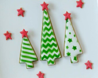 May include: A white plate displays three Christmas tree-shaped cookies with green and white icing designs, topped with red star sprinkles. Additional red star sprinkles are scattered around the cookies, creating a festive holiday scene.