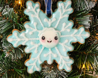 Könnte beinhalten: Ein Lebkuchen-Schneeflocken-Weihnachtsschmuck, verziert mit hellblauer Glasur und weißen Details, mit einem lächelnden Gesicht. Er hängt an einem blauen Band vor einem Weihnachtsbaumhintergrund. Der Durchmesser beträgt ca. 10 cm.
