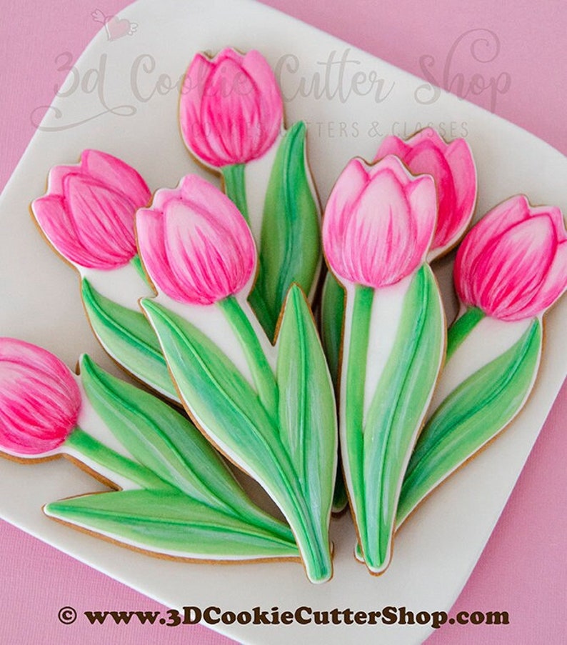 May include: A bouquet of pink and green sugar cookies decorated to look like tulips. The cookies are arranged on a white plate with a pink background. The text "3D Cookie Cutter Shop" is visible in the top left corner of the image.