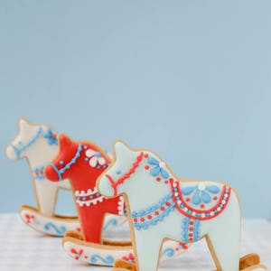 May include: Three decorated rocking horse cookies in a row. The cookies are light blue, red, and white, with intricate icing details in red and blue. The cookies are on a light blue and white checkered surface.