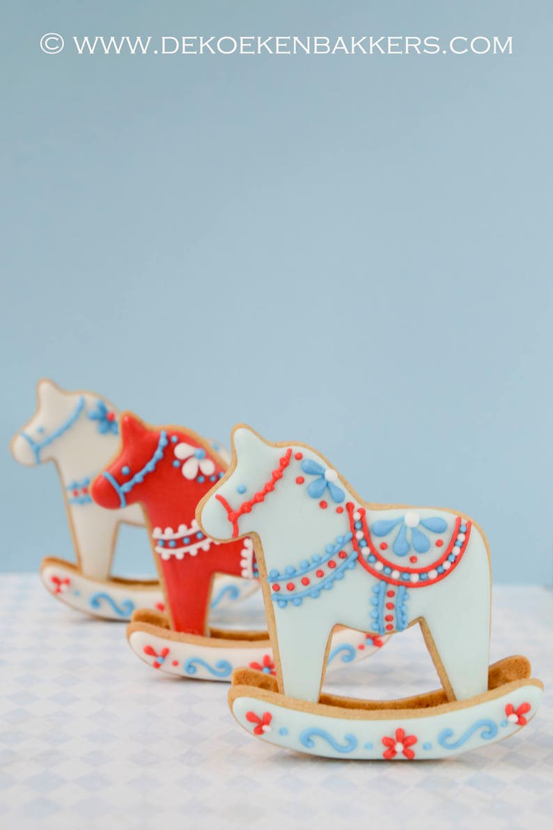 May include: Three decorated rocking horse cookies in a row. The cookies are light blue, red, and white, with intricate icing details in red and blue. The cookies are on a light blue and white checkered surface.