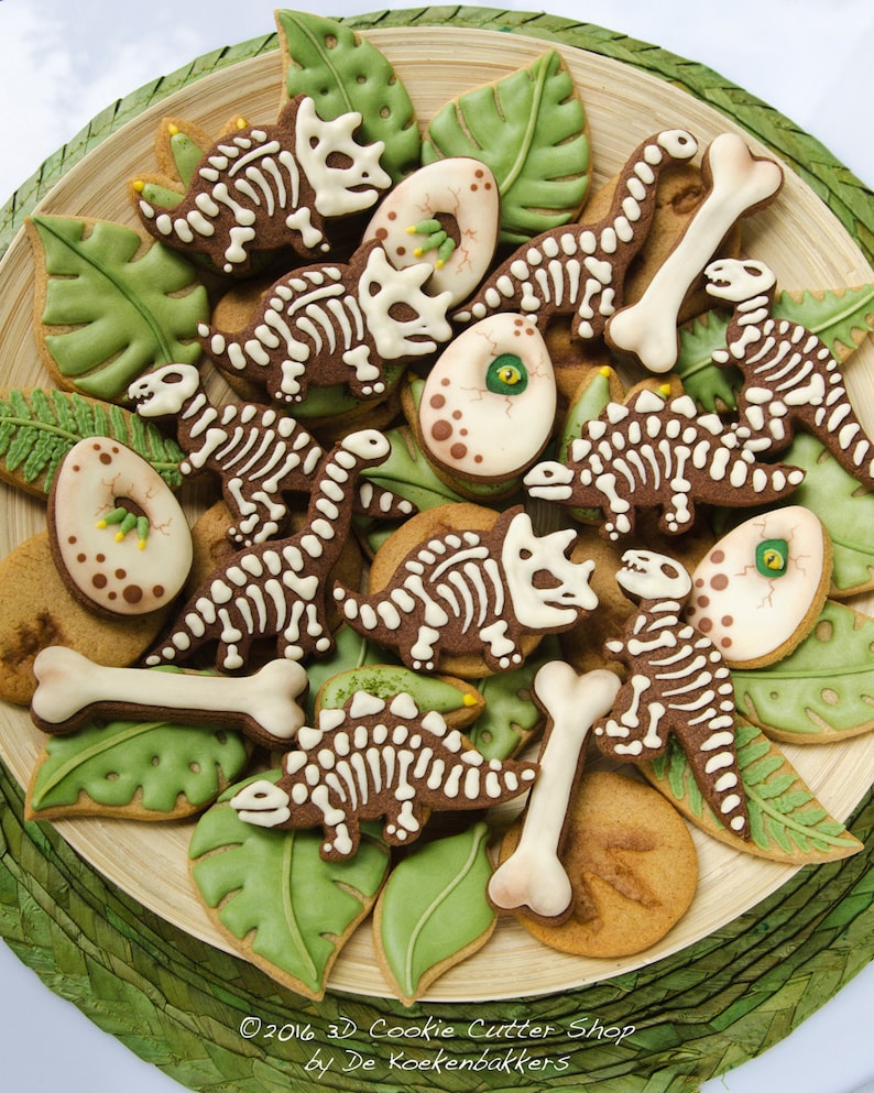 May include: A wooden plate filled with dinosaur-themed cookies. The cookies are decorated with brown icing and white bone details, along with green leaf-shaped cookies and egg-shaped cookies. The cookies are arranged on a green woven mat.