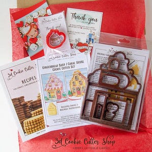 May include: A collection of cookie cutters and related items, including a gingerbread shop/canal house cookie cutter set, recipe cards, and thank you notes. The cutters are brown, and the packaging includes red and white accents.
