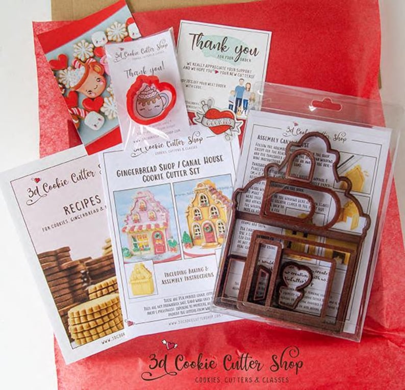 May include: A collection of cookie cutters and related items, including a gingerbread shop/canal house cookie cutter set, recipe cards, and thank you notes. The cutters are brown, and the packaging includes red and white accents.