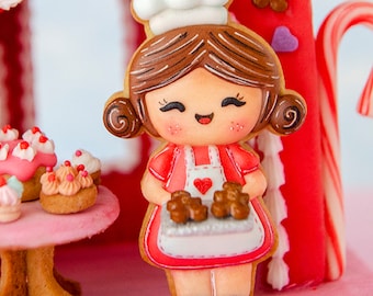 May include: A gingerbread cookie in the shape of a smiling baker girl wearing a chef's hat and apron, holding two gingerbread men. The cookie is set against a red gingerbread house with candy cane accents and cupcakes on a pink table.