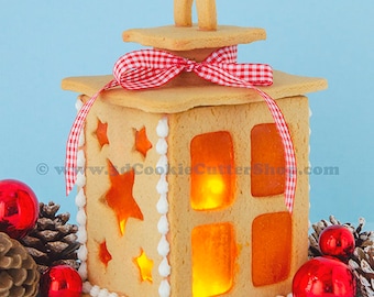 May include: A gingerbread lantern decorated with white icing and red and white gingham ribbon. The lantern has a light inside and is surrounded by pine cones and red ornaments.