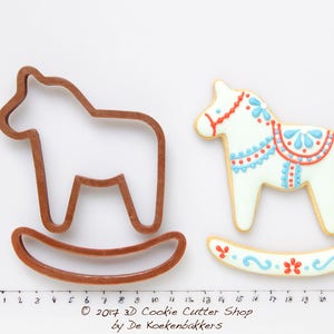 May include: A brown rocking horse cookie cutter next to a decorated cookie. The cookie is white with red and blue details. The rocking horse cookie cutter is made of brown plastic. The cookie is decorated with red and blue floral designs.