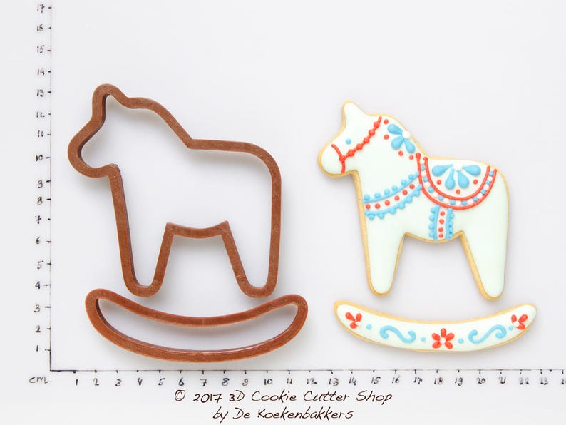 May include: A brown rocking horse cookie cutter next to a decorated cookie. The cookie is white with red and blue details. The rocking horse cookie cutter is made of brown plastic. The cookie is decorated with red and blue floral designs.