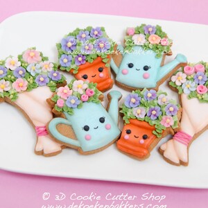 Op de afbeelding: Vijf gedecoreerde suikerkoekjes in de vorm van een gieter, een bloempot en een boeket bloemen. De koekjes zijn versierd met pastelkleuren en hebben schattige gezichtjes. De koekjes liggen op een wit bord met een roze achtergrond. © 3D Cookie Cutter Shop www.dekoekenbakkers.com
