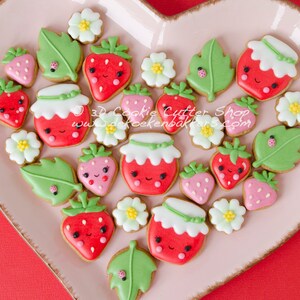 May include: A pink heart-shaped plate filled with decorated sugar cookies. The cookies are shaped like strawberries, leaves, and jars of jam. The cookies have cute faces and are decorated with red, green, and white icing. The plate is on a red background. 3D Cookie Cutter Shop www.dekoekenbakkers.com