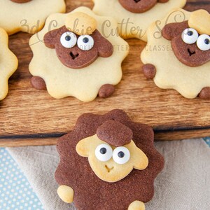 May include: A close-up of five decorated sugar cookies shaped like sheep. The cookies are brown and tan with white icing eyes and a brown nose. The cookies are on a wooden surface. The text "3d Cookie Cutter Shop" is visible in the image.