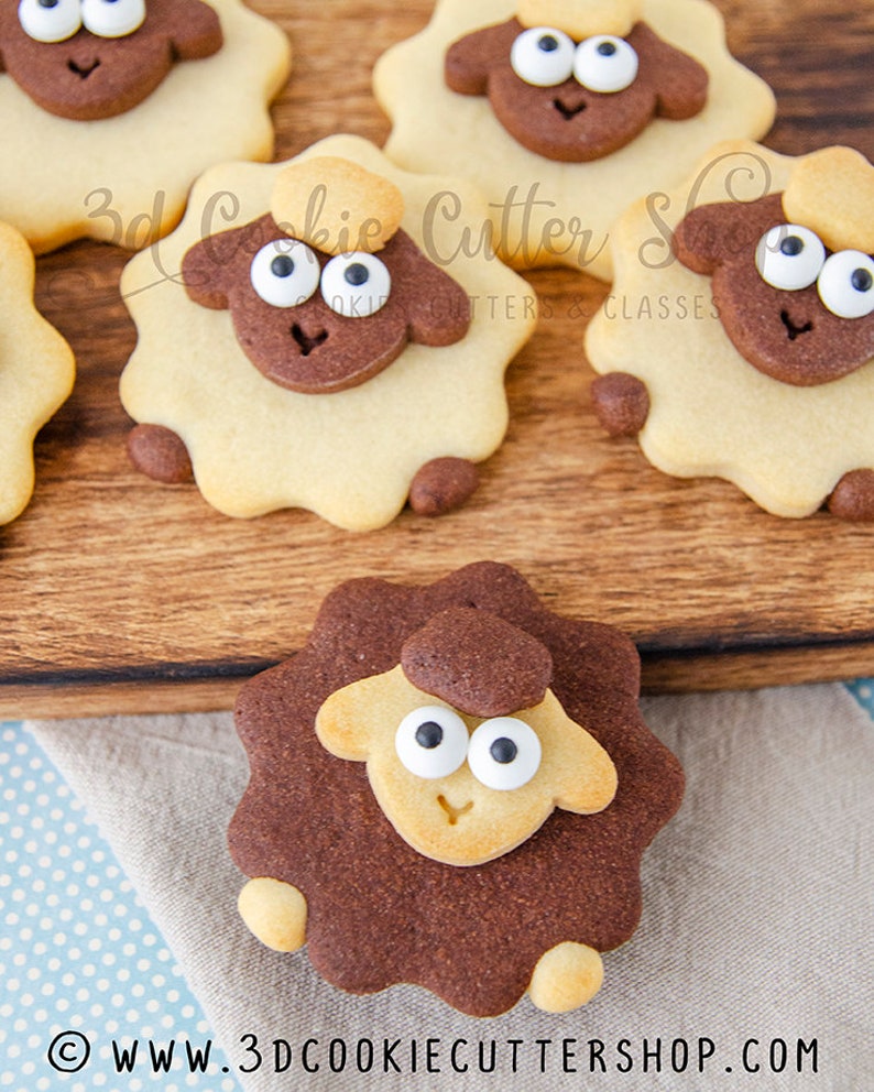 May include: A close-up of five decorated sugar cookies shaped like sheep. The cookies are brown and tan with white icing eyes and a brown nose. The cookies are on a wooden surface. The text "3d Cookie Cutter Shop" is visible in the image.