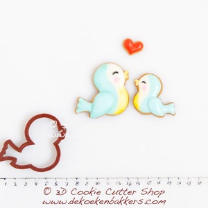 May include: Two blue and yellow bird cookie cutters with a red heart cookie cutter. The cookie cutters are on a white surface with a ruler in centimeters.