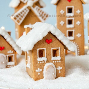 Puede incluir: Un pueblo de jengibre con un techo cubierto de nieve, un molino de viento y una casa alta de jengibre con corazones rojos. Las casas están decoradas con glaseado blanco y chispas. El pueblo está sentado sobre una superficie blanca que parece nieve.