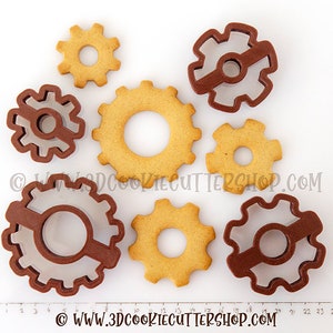 May include: A set of eight cookie cutters shaped like gears. The cutters are made of brown plastic and have a variety of sizes. The cutters are arranged on a white surface with a ruler in the background.