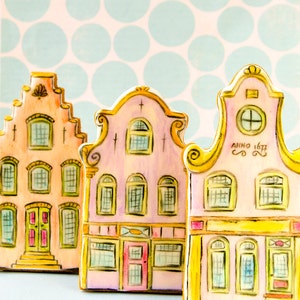 Puede incluir: Tres cortadores de galletas pintados a mano con forma de casas holandesas. Las casas están pintadas en colores pastel con detalles como ventanas, puertas y techos. Los cortadores de galletas están sobre un fondo azul claro con lunares blancos.