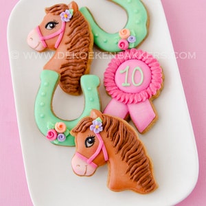 Può includere: Quattro biscotti di zucchero decorati su un piatto bianco. Due biscotti a forma di testa di cavallo marroni, un biscotto a forma di ferro di cavallo verde e un biscotto rosa con un nastro bianco e il numero 10.
