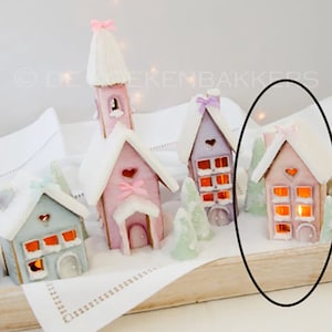 Può includere: Una collezione di cinque case di pan di zenzero in miniatura decorate con glassa rosa pastello, blu e viola. Le case hanno tetti bianchi e finestre con luci arancioni incandescenti all'interno. Le case sono disposte su un vassoio di legno con una tovaglia bianca sotto.