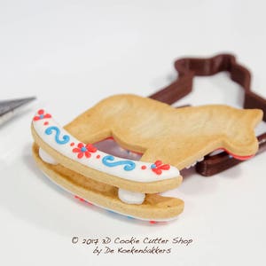 May include: A decorated rocking horse cookie with red and blue floral accents, next to a brown cookie cutter. A pastry bag is in the background. The cookie is decorated with white icing and blue swirls. The text "&copy; 2017 3D Cookie Cutter Shop by De Koekenbakkers" is visible.