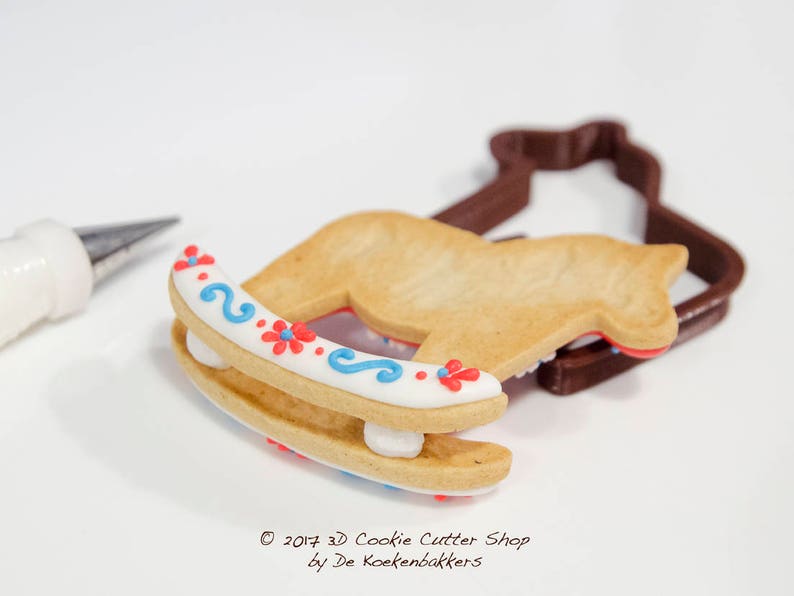 May include: A decorated rocking horse cookie with red and blue floral accents, next to a brown cookie cutter. A pastry bag is in the background. The cookie is decorated with white icing and blue swirls. The text "&copy; 2017 3D Cookie Cutter Shop by De Koekenbakkers" is visible.