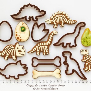 May include: A collection of dinosaur-themed cookies and cookie cutters. The cookies include dinosaur skeletons, footprints, and an egg. The cookie cutters are brown, and the cookies are decorated with white and brown icing. The image includes the text "&copy;2016 3D Cookie Cutter Shop by De Koekenbakkers."
