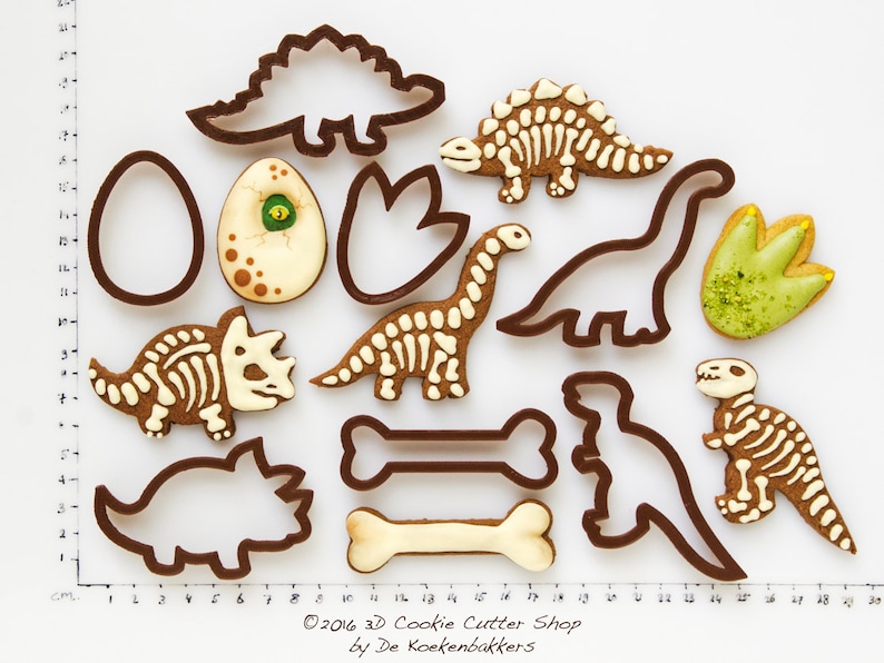May include: A collection of dinosaur-themed cookies and cookie cutters. The cookies include dinosaur skeletons, footprints, and an egg. The cookie cutters are brown, and the cookies are decorated with white and brown icing. The image includes the text "&copy;2016 3D Cookie Cutter Shop by De Koekenbakkers."