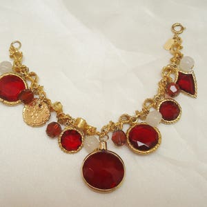 May include: A gold charm bracelet with red and white glass beads. The bracelet features a variety of charms, including coins, hearts, and other shapes.