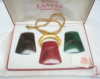 May include: A gold chain necklace with a red, green, and brown stone pendant set in a gold frame. The necklace is in a white box with a red lining and a Lanvin label. The box lid has the text "Bijoux Lanvin New York-Paris".