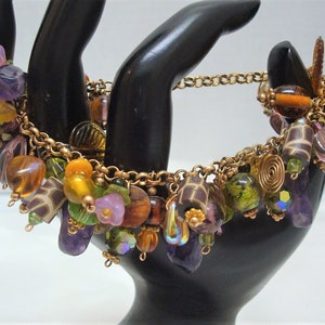 May include: A gold chain bracelet with a variety of colorful beads, including purple, pink, green, yellow, and brown. The beads are made of glass, metal, and wood.