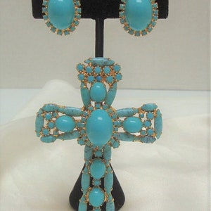 May include: A turquoise and gold cross pendant with a large turquoise stone in the centre. The pendant is surrounded by smaller turquoise stones and gold accents. The pendant is on a black stand.