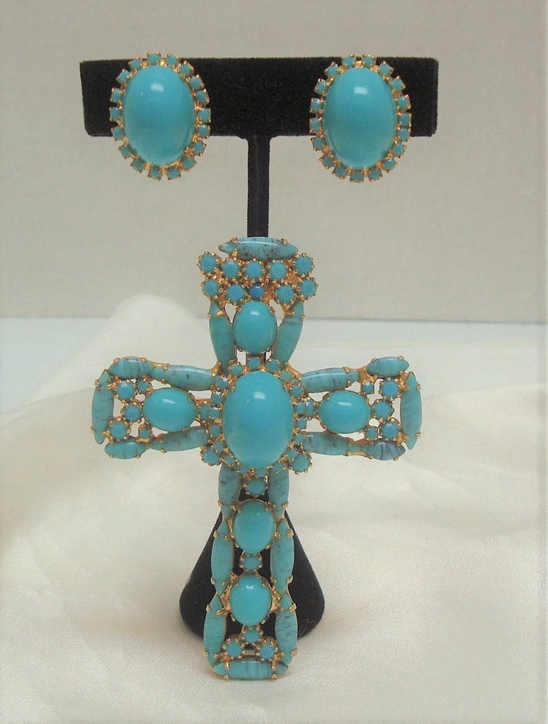 May include: A turquoise and gold cross pendant with a large turquoise stone in the centre. The pendant is surrounded by smaller turquoise stones and gold accents. The pendant is on a black stand.