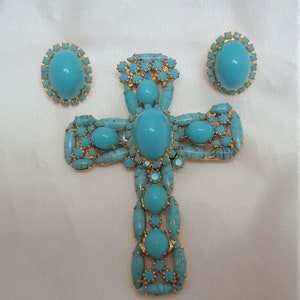 May include: A turquoise and gold cross pendant and earrings set. The cross is large and features a central turquoise stone surrounded by smaller turquoise stones. The earrings are small and feature a single turquoise stone.