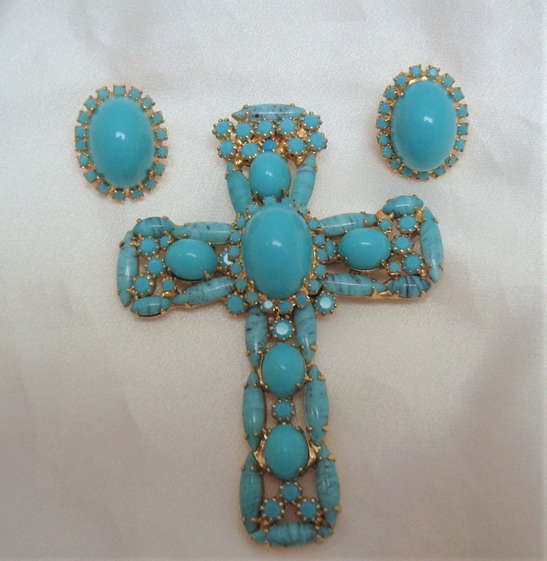 May include: A turquoise and gold cross pendant and earrings set. The cross is large and features a central turquoise stone surrounded by smaller turquoise stones. The earrings are small and feature a single turquoise stone.