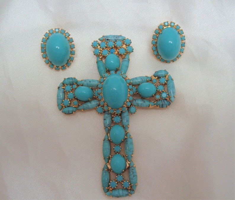 May include: A turquoise blue cross pendant with matching earrings. The cross is made of gold-toned metal and is adorned with turquoise blue stones.