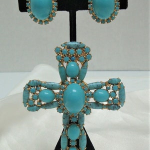 May include: A turquoise and gold cross pendant necklace with matching earrings. The cross is made of metal and has a large turquoise stone in the centre. The earrings are also made of metal and have a smaller turquoise stone in the centre.