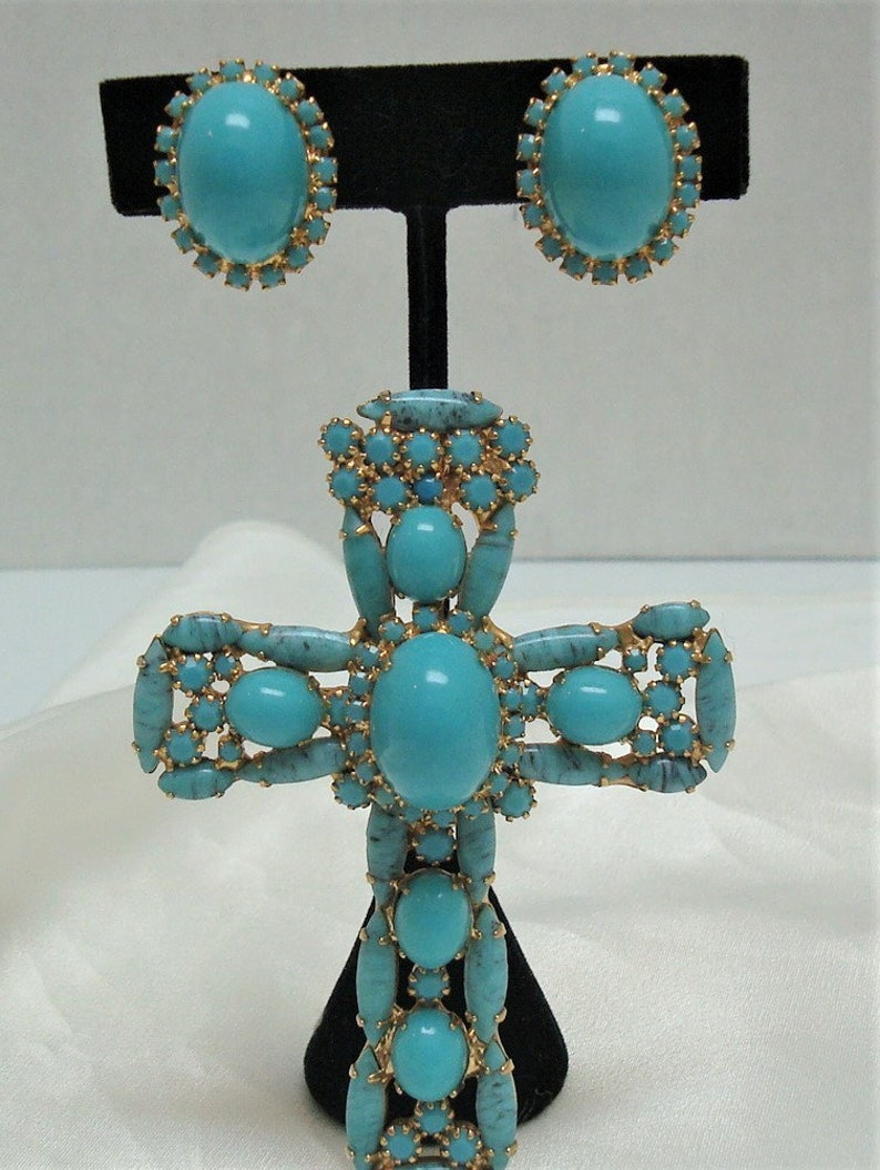 May include: A turquoise and gold cross pendant necklace with matching earrings. The cross is made of metal and has a large turquoise stone in the centre. The earrings are also made of metal and have a smaller turquoise stone in the centre.