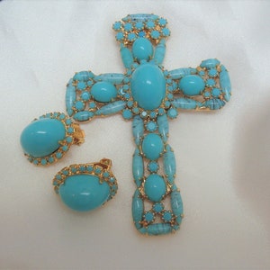 May include: A turquoise and gold cross pendant necklace and matching earrings. The cross is made of gold-tone metal and features a large turquoise stone in the centre. The earrings are also gold-tone metal and feature smaller turquoise stones.