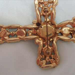 May include: Gold tone cross brooch with clear and blue rhinestones. The back of the brooch has a pin and a signature that reads "Alice Caviness".